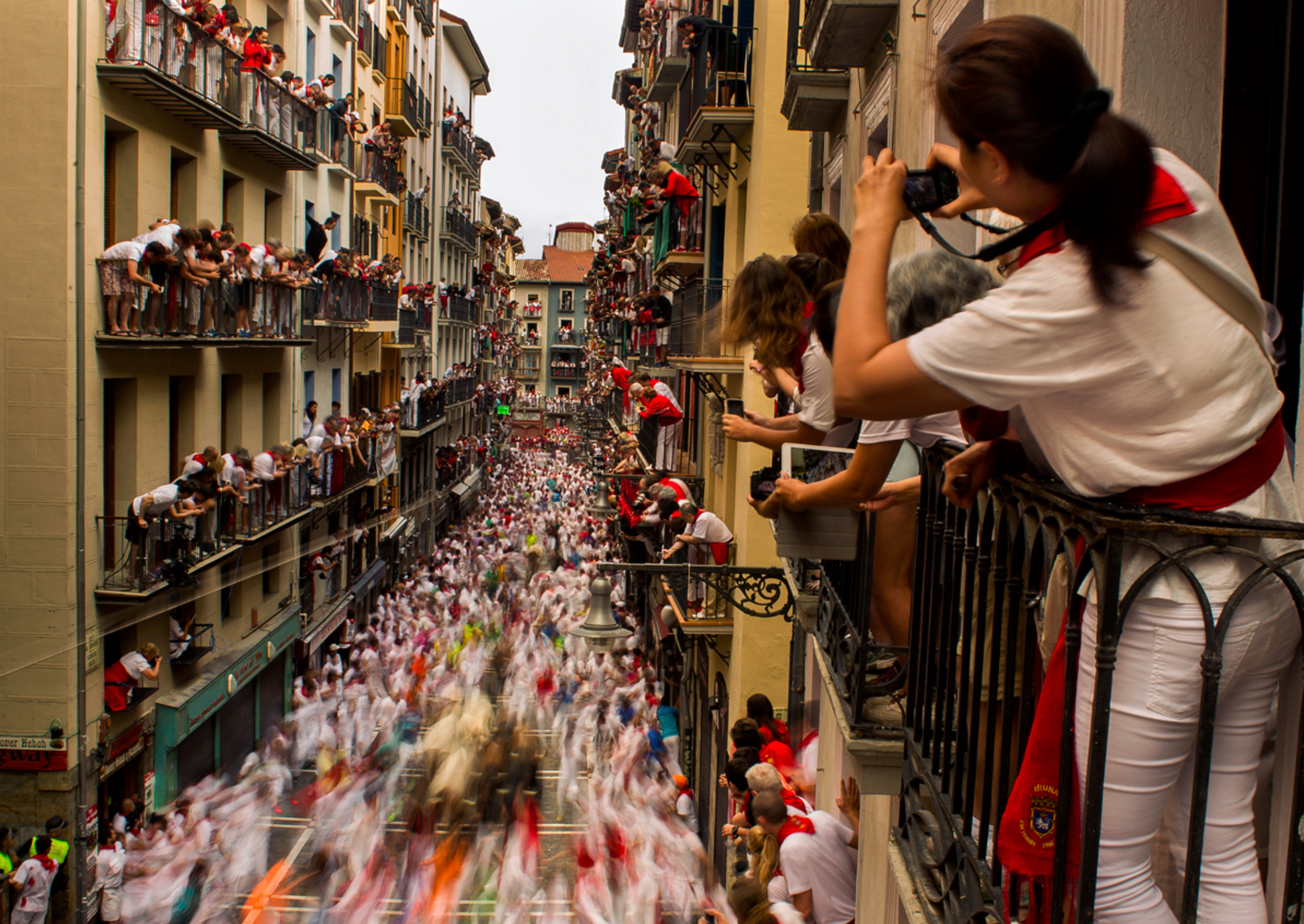 Balconies full of spectators on Calle Estafeta (Estafeta Street) watch the Pamplona bull run. Bulls and runners in the street below are blurry, capturing their speed. 